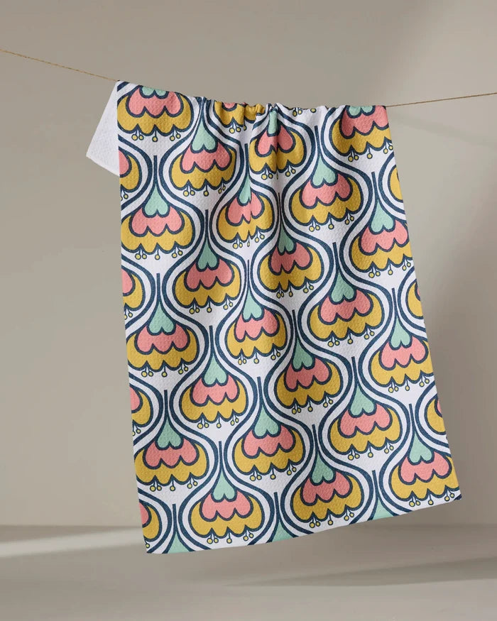 The Renee Fly "70s Floral" recycled kitchen towel, featuring vibrant retro shapes in yellow, orange, pink, and blue, hangs on a string against a neutral background.