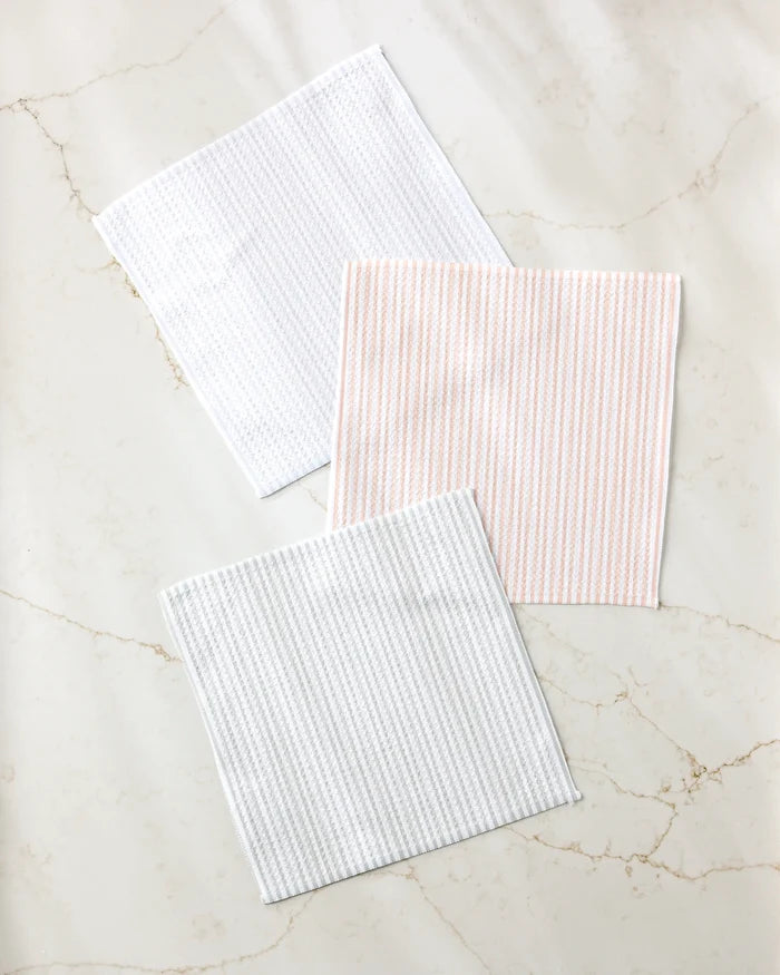 Three square À La Carte cloth napkins by Kailee Wright, made from recycled materials, are displayed on a light marble surface: one plain white, one with light pink stripes, and one with light gray stripes.