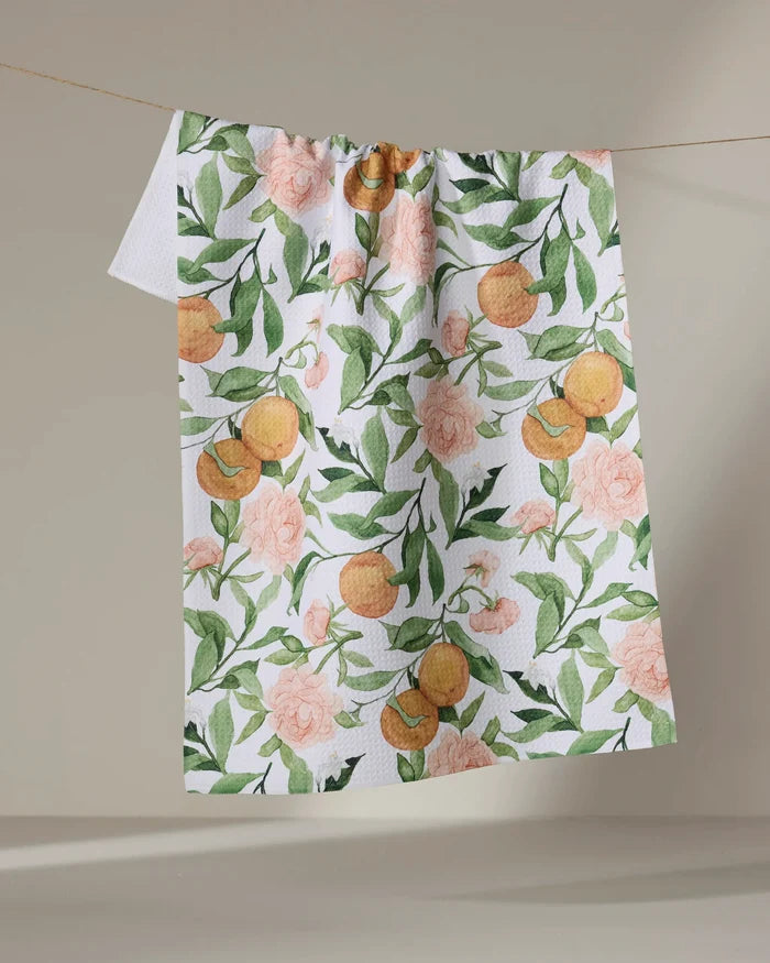 The "A Peony For Your Thoughts" recycled kitchen towel by Aubrey Fairchild, with a vibrant floral and foliage pattern, hangs on a line against a plain beige background.