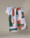 The Abstract Collage recycled kitchen towel by Claire Iglesias, featuring geometric shapes in green, rust, white, and lavender, hangs on a line against a neutral background.