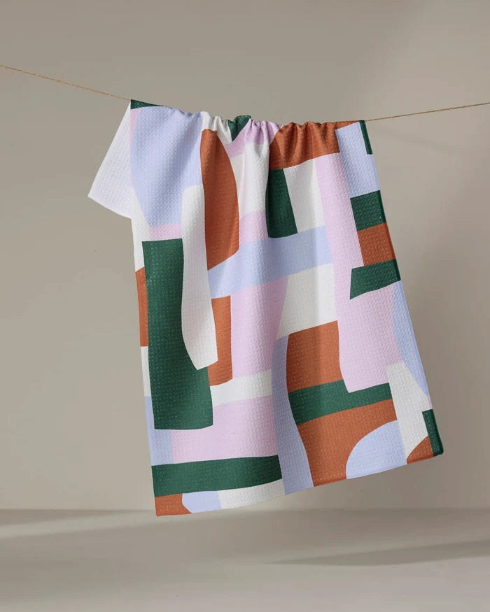 The Abstract Collage recycled kitchen towel by Claire Iglesias, featuring geometric shapes in green, rust, white, and lavender, hangs on a line against a neutral background.