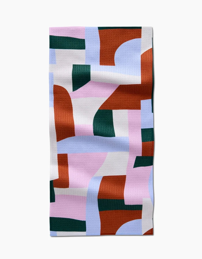 The Abstract Collage bar towel by Claire Iglesias is rectangular, made from recycled materials, and features an abstract pattern with rust orange, forest green, light blue, white, and pink shapes on a textured surface.