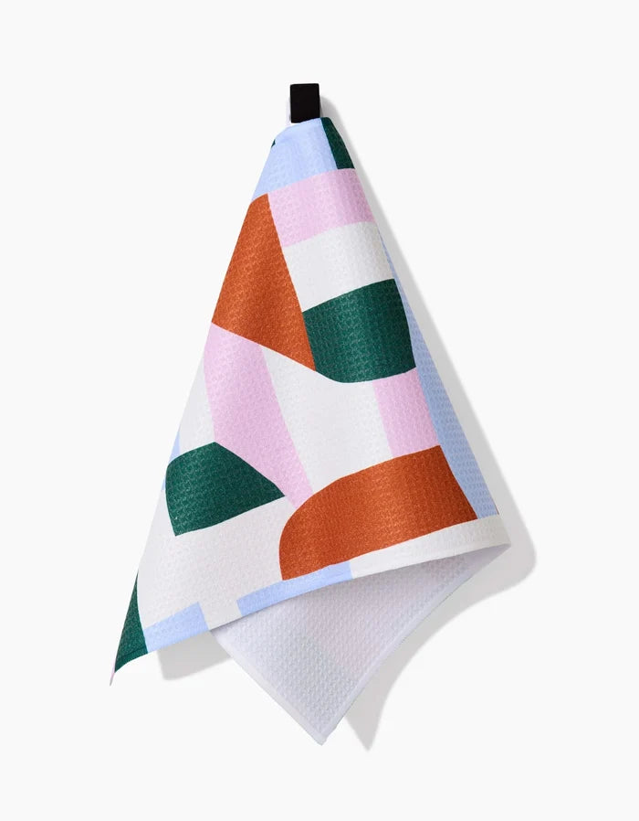 The Abstract Collage bar towel by Claire Iglesias features a geometric pattern in orange, green, pink, and white Geoweave fabric with a black hanging loop. The towel casts a shadow on the white background.
