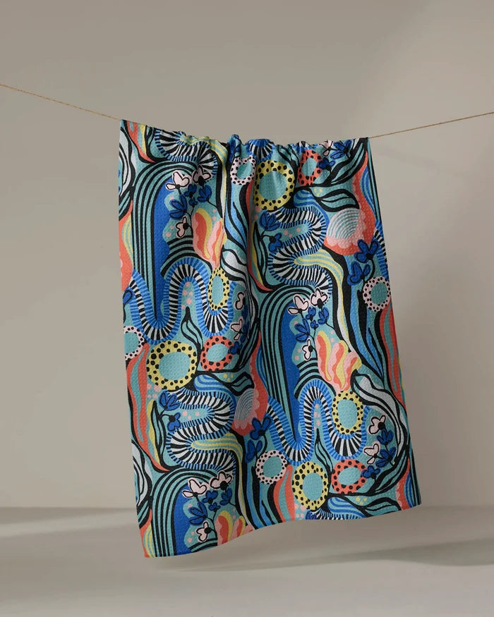The "Abstract Dreams" kitchen towel by Masha Volnova features bold wavy blue and green lines, vibrant red, pink, and yellow shapes, plus scattered florals. It hangs from a string against a neutral background.