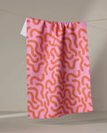 The Abstract Snake kitchen towel by Jennifer Bouron, featuring a pink and orange wavy design, hangs on a string against a neutral background.