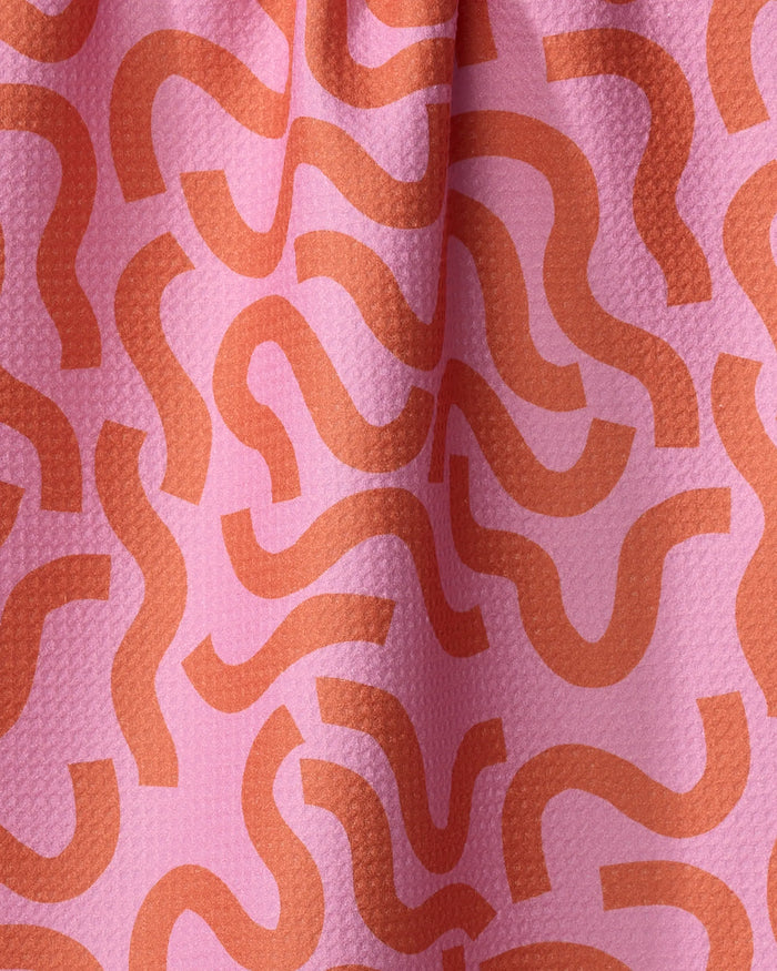 A close-up of the Jennifer Bouron "Abstract Snake" kitchen towel shows pink recycled fabric with an orange, wavy pattern, a slightly bumpy texture, and gentle folds.