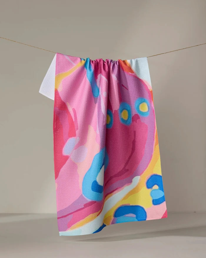 The "Abundance" towel by Sareka Unique, featuring quick-drying fabric and vibrant abstract patterns in pink, blue, yellow, and white, hangs on a string against a neutral beige background.
