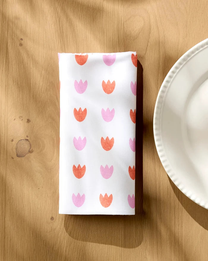 A folded white Addison dinner napkin by Amy MacCready, featuring pink and orange tulips, sits on a wooden table beside a scalloped plate. Sunlight and subtle shadows highlight these absorbent napkins.
