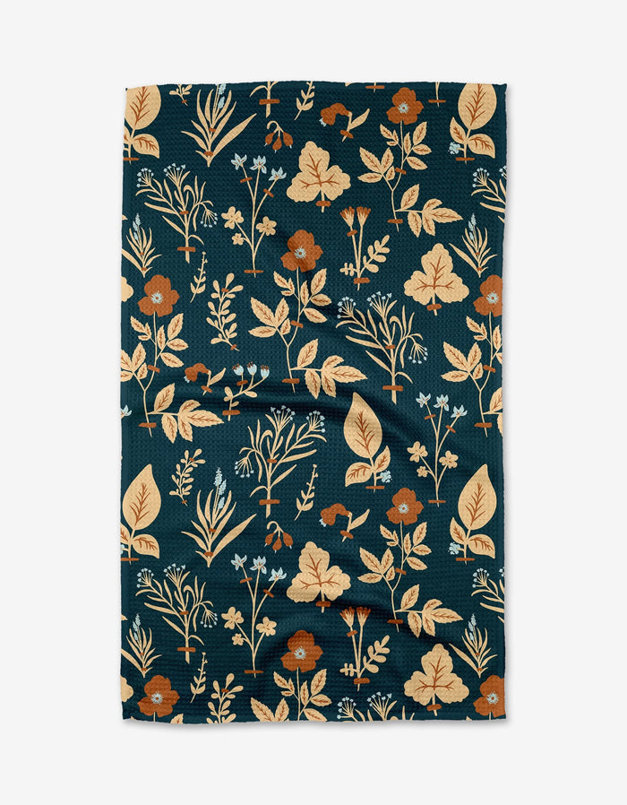 The Aemilius tea towel by Danelys Sidron features a dark green Geoweave fabric with beige and orange floral and leaf patterns for a botanical touch, shown slightly wrinkled against a white background.