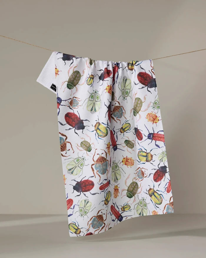 The "African Beetles" recycled kitchen towel by Taryn Brennan, featuring colorful beetle illustrations, hangs on a clothesline against a neutral backdrop.