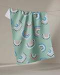 The "After the Rain" tea towel by Claire Iglesias features a light green textured fabric with colorful pink, white, blue, and black semi-circles and arches, hanging on a line against a neutral background.