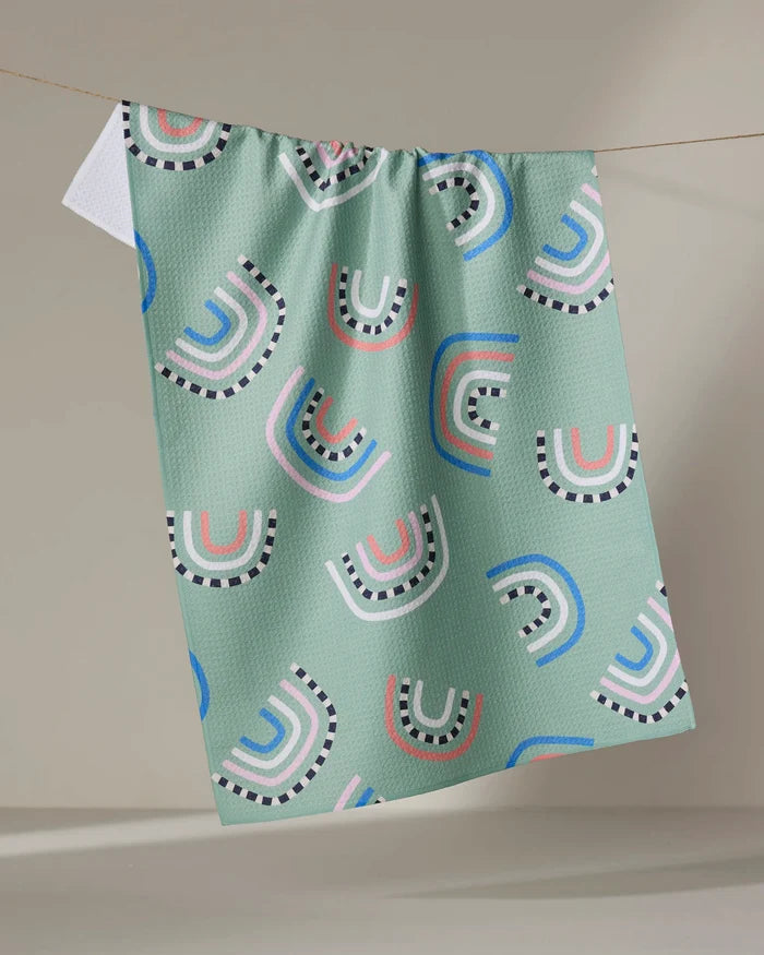 The "After the Rain" tea towel by Claire Iglesias features a light green textured fabric with colorful pink, white, blue, and black semi-circles and arches, hanging on a line against a neutral background.