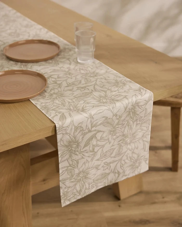 The Alba table by Geometry features a stain-resistant, beige floral table runner, two stacked beige ceramic plates, and two clear glasses. A wooden chair is partially visible beside the light wooden table.