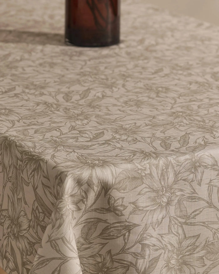 A close-up of the Geometry Alba tablecloth, beige and stain-resistant with a subtle floral pattern, covers the table. The bottom of a dark glass vase appears at the images top.