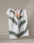 The "Along the Path" recycled kitchen towel by Elana Gabrielle, featuring a jagged abstract green and peach floral design, hangs on a clothesline against a neutral background.