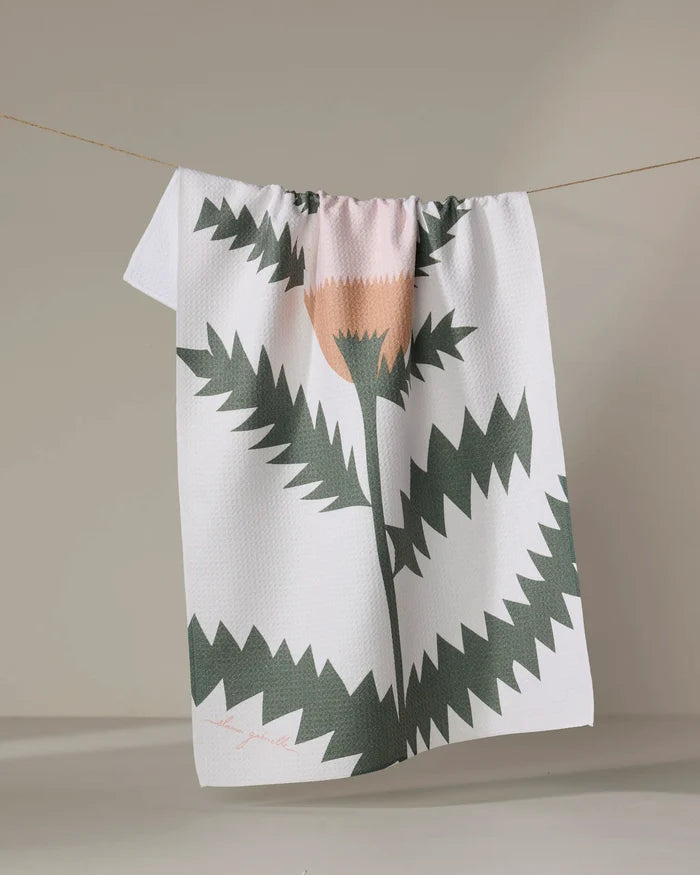 The "Along the Path" recycled kitchen towel by Elana Gabrielle, featuring a jagged abstract green and peach floral design, hangs on a clothesline against a neutral background.