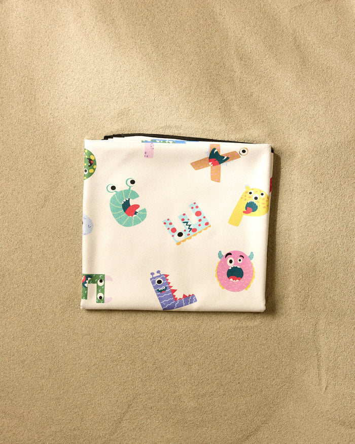 The Vaishali Design Studio "Alphabet Monsters" beach blanket features colorful cartoon letters and monster faces on a light background, displayed folded on a beige textured surface.