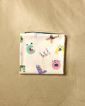 The Vaishali Design Studio "Alphabet Monsters" beach blanket features colorful cartoon letters and monster faces on a light background, displayed folded on a beige textured surface.