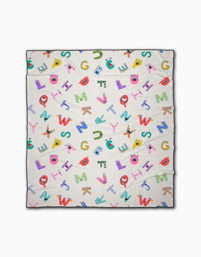 The Alphabet Monsters beach blanket by Vaishali Design Studio features playful cartoon alphabet letters with fun faces on a cream background. Made from quick-drying recycled materials, it offers both eco-friendly comfort and vibrant style.