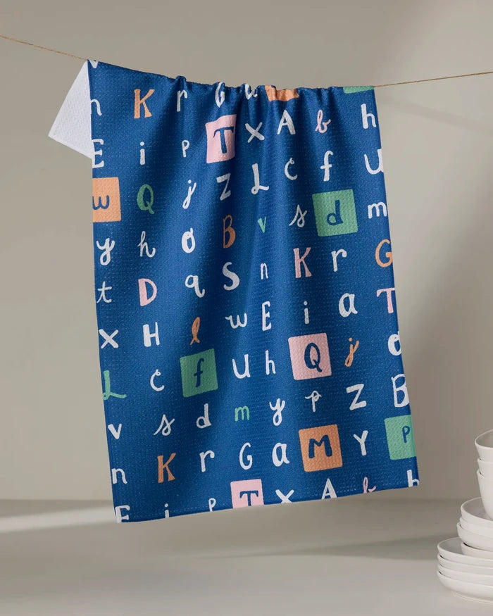 The 'Alphabet' tea towel by Amy MacCready is a blue, recycled design with vibrant, varied letters—some boxed. Quick-drying and hanging on a line, it’s pictured near stacked white dishes on a light surface.