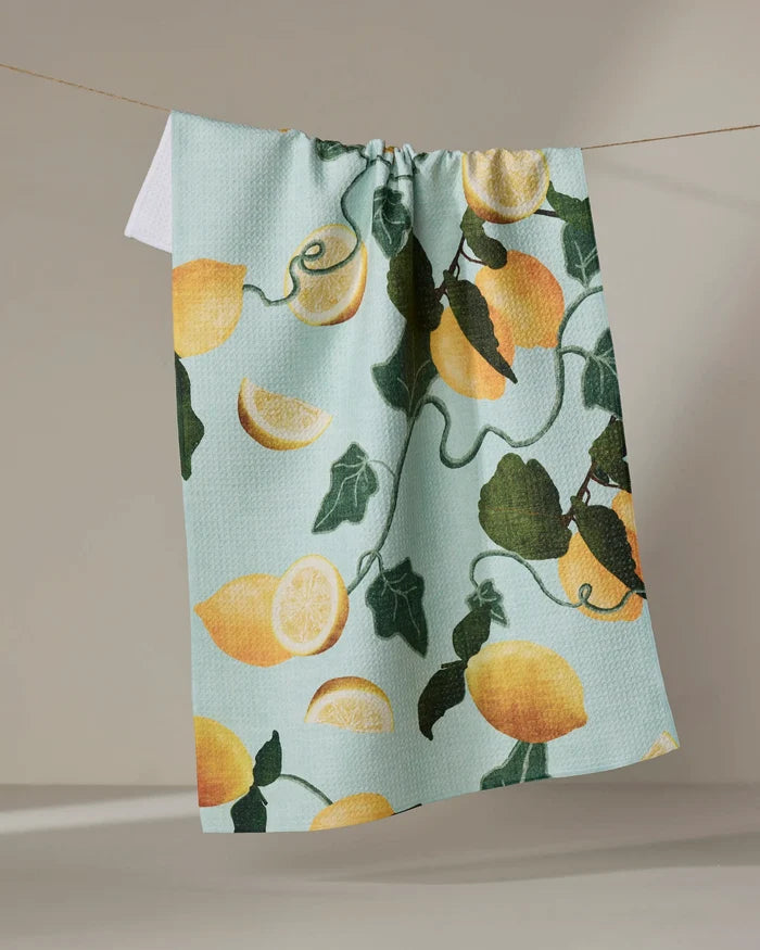 The Pascale Dilger "Amalfi Lemons" tea towel features whole lemons, slices, and green leaves on light blue, hanging on a clothesline. Made from quick-drying recycled materials, it’s an eco-friendly and stylish choice.