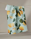 The Pascale Dilger "Amalfi Lemons" tea towel features whole lemons, slices, and green leaves on light blue, hanging on a clothesline. Made from quick-drying recycled materials, it’s an eco-friendly and stylish choice.