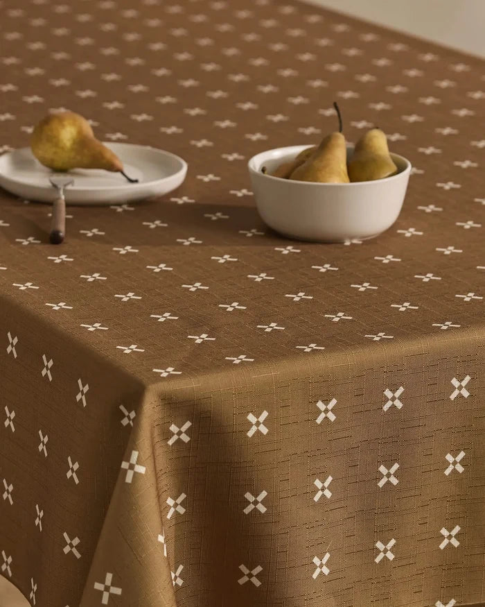 The table is covered with the Amber Stitch tablecloth by Geometry, featuring a white X pattern. A white plate with a pear and a bowl with three pears sit on top, set against a neutral, minimal background.