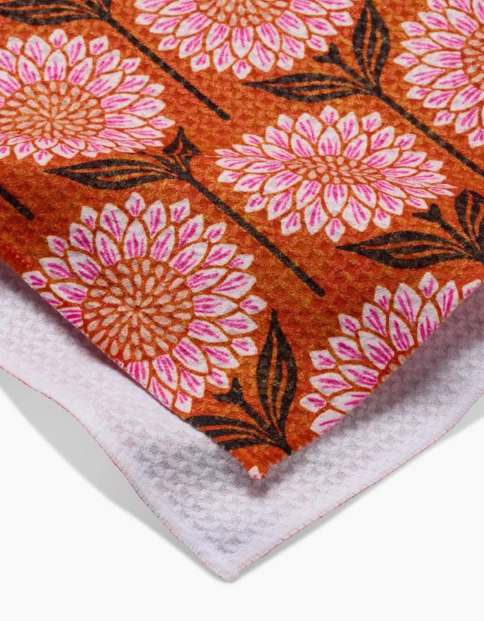 The Amber tea towel by Amy MacCready features an orange top with large pink and white floral designs, dark brown stems and leaves, a textured white underside, and is crafted from ultra-absorbent recycled materials.