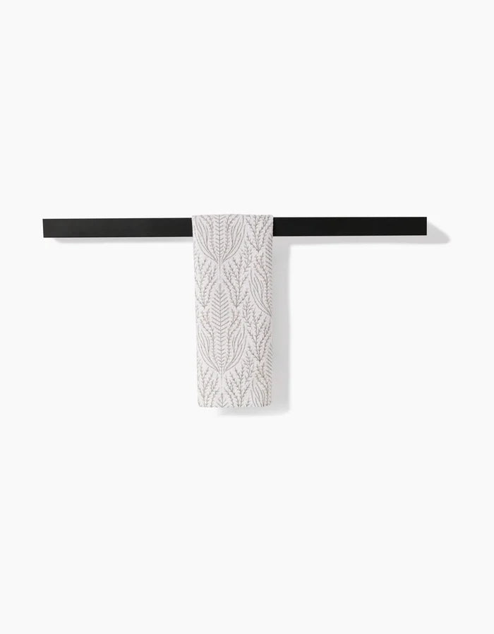 The Amberlin bar towel by Amy MacCready, featuring a subtle leaf pattern and made from ultra-absorbent recycled materials, hangs on a minimalist black towel bar against a white wall.