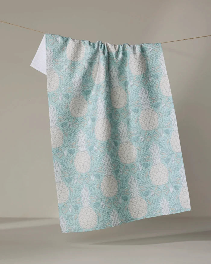 The Andre tea towel by Amy MacCready, featuring a light blue and white pineapple pattern and made from recycled materials, hangs over a clothesline against a plain, neutral background.