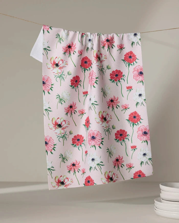 The Linda Fox Anemone Garden Flowers pink tea towel, featuring a colorful floral pattern, hangs on a clothesline with white plates nearby on a neutral surface.