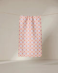 The GEOMETRY Anew towel features ultra-absorbent fabric and a geometric pattern of intersecting pink and orange lines forming diamonds, shown hanging on a line against a neutral, light background.