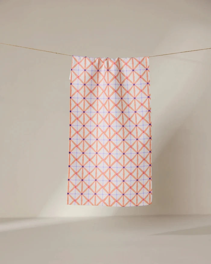 The GEOMETRY Anew towel features ultra-absorbent fabric and a geometric pattern of intersecting pink and orange lines forming diamonds, shown hanging on a line against a neutral, light background.