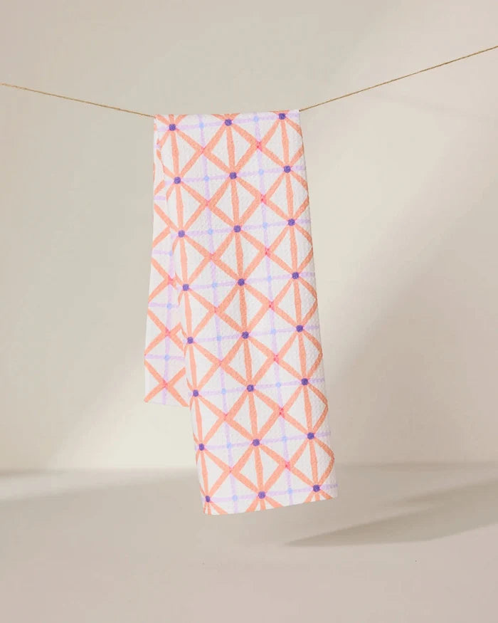 The GEOMETRY Anew tea towel features a quick-drying design with a pink and orange geometric crisscross pattern and small purple dots, shown hanging by a string against a neutral background.