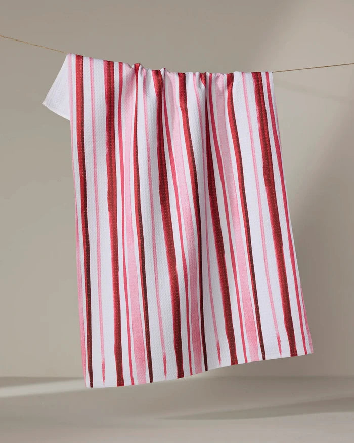 The Aperol in Rome Stripe kitchen towel by Travel Write Draw by Meagan Morrison, featuring recycled fabric and vertical red and pink stripes, hangs on a clothesline against a neutral background.