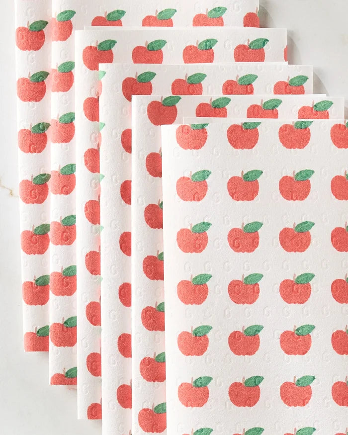 Brittany Jepsen’s Apple Grove features six white reusable paper towels stacked in a staggered pile, each adorned with a repeating red apple and green leaf pattern on a light, neutral surface.