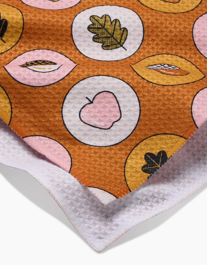 A close-up of Megan Roys Apples Acorns recycled kitchen towel shows a brown, pink, and yellow leaf and apple pattern in circles with a white, waffle-textured edge.