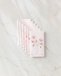 Seven packets of Linda Fox's April Sweet Pea seeds, featuring sweet pea flower illustrations, are fanned out on white marble beside a stack of eco-friendly kitchen towels for a fresh and sustainable vibe.