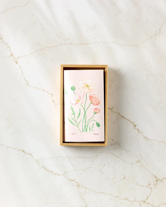 A rectangular box of Linda Fox April Sweet Pea eco-friendly kitchen towels, featuring a floral design of pink and yellow poppies on the lid, rests on a light marble surface as sunlight casts gentle shadows.