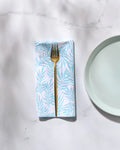 A gold fork rests on a folded, white washable GEOMETRY "Aqua Branches" napkin with a light blue leaf pattern, next to a light blue plate on a white marble surface.