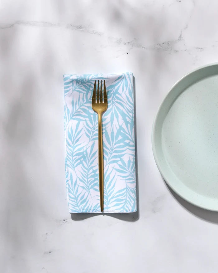 A gold fork rests on a folded, white washable GEOMETRY "Aqua Branches" napkin with a light blue leaf pattern, next to a light blue plate on a white marble surface.