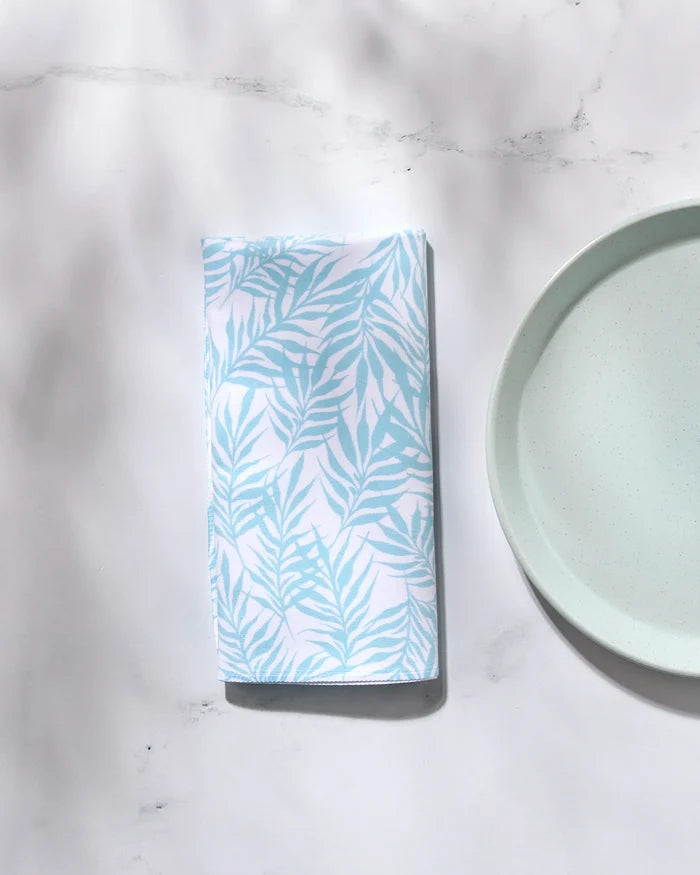 The GEOMETRY Aqua Branches washable cloth napkin with a light blue leaf pattern is folded beside a pale blue plate on a white marble surface with soft shadows.