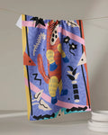 The Ariranha tea towel by Camila Leão, made from recycled materials, showcases a vibrant abstract design with a playful animal and bold patterns, hanging above white plates.