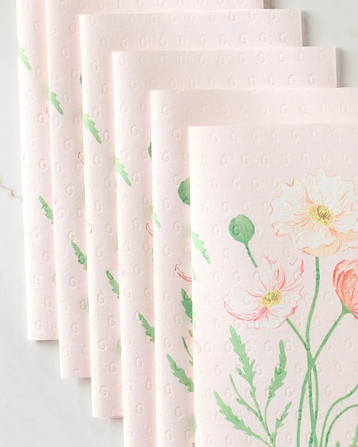 Six "August Poppy" napkins by Linda Fox, crafted from eco-friendly paper, are fanned out to display their delicate poppy floral print in soft green, peach, and yellow on a textured pastel pink surface.