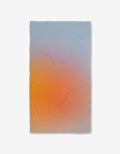 The GEOMETRY Aura towel is a rectangular Luxe Bath Towel with a gradient from soft blue to warm orange and yellow, made with ultra-absorbent Geoweave technology. Shown on a white background.