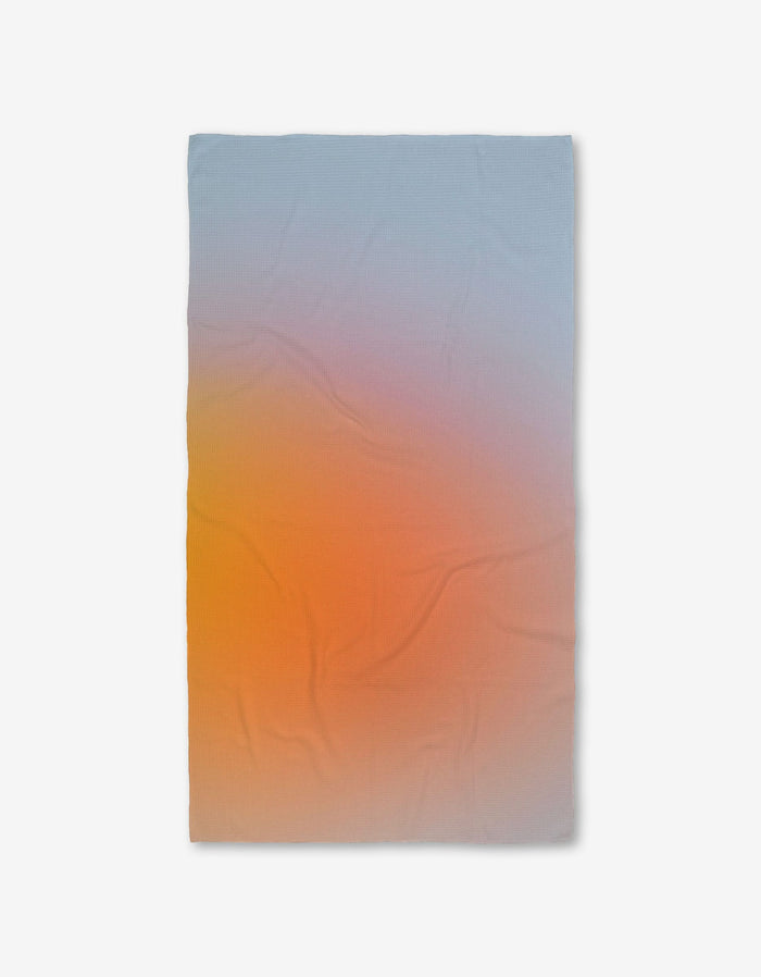 The GEOMETRY Aura towel is a rectangular Luxe Bath Towel with a gradient from soft blue to warm orange and yellow, made with ultra-absorbent Geoweave technology. Shown on a white background.