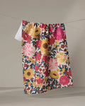 The "Autumn Blooms" tea towel by Rebecca Bobko, featuring a vibrant floral design in red, pink, yellow, and green, hangs from a thin string against a plain, neutral background.