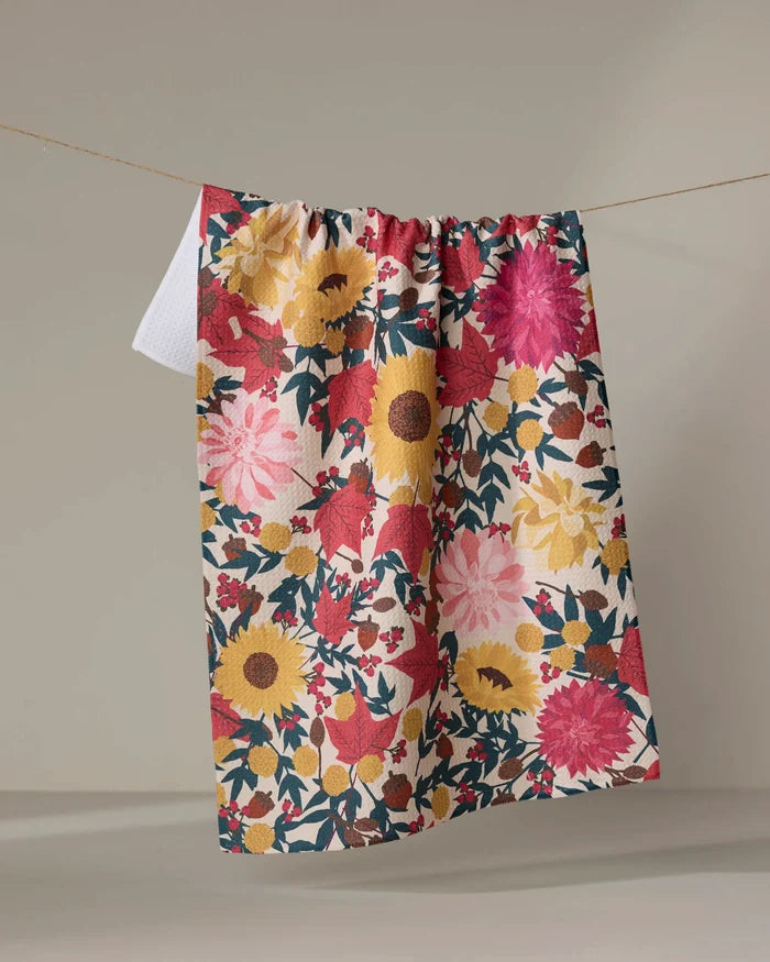The "Autumn Blooms" tea towel by Rebecca Bobko, featuring a vibrant floral design in red, pink, yellow, and green, hangs from a thin string against a plain, neutral background.