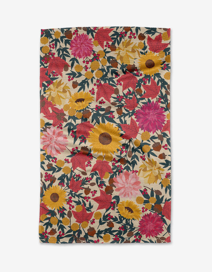 The Rebecca Bobko Autumn Blooms Geoweave™ tea towel features a vibrant floral pattern of red, pink, and yellow flowers with green leaves and berries on a cream background. The rectangular fabric has a slightly wrinkled appearance.
