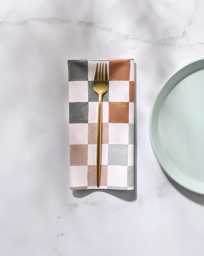 A gold fork rests on the folded GEOMETRY Autumn Checkers reusable napkin, featuring gray, tan, and white squares, placed beside a light blue plate on a white marble surface.